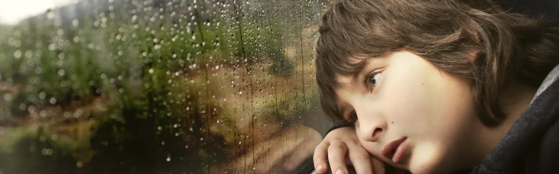 Niño pensativo mirando por una ventana con gotas de lluvia.